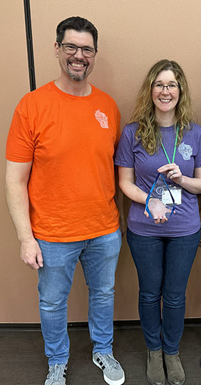 A man with dark hair and a beard wearing an orange shirt and jeans stands to the left of a woman with long brown hair wearing a purple shirt and jeans and holding a clear crystal award.