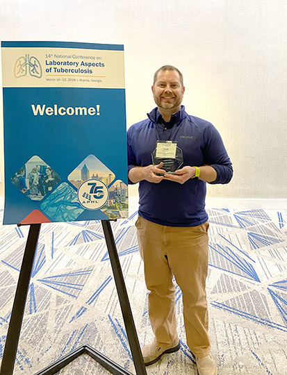 A man with a beard wearing a blue shirt and tan pants holds a clear crystal award.